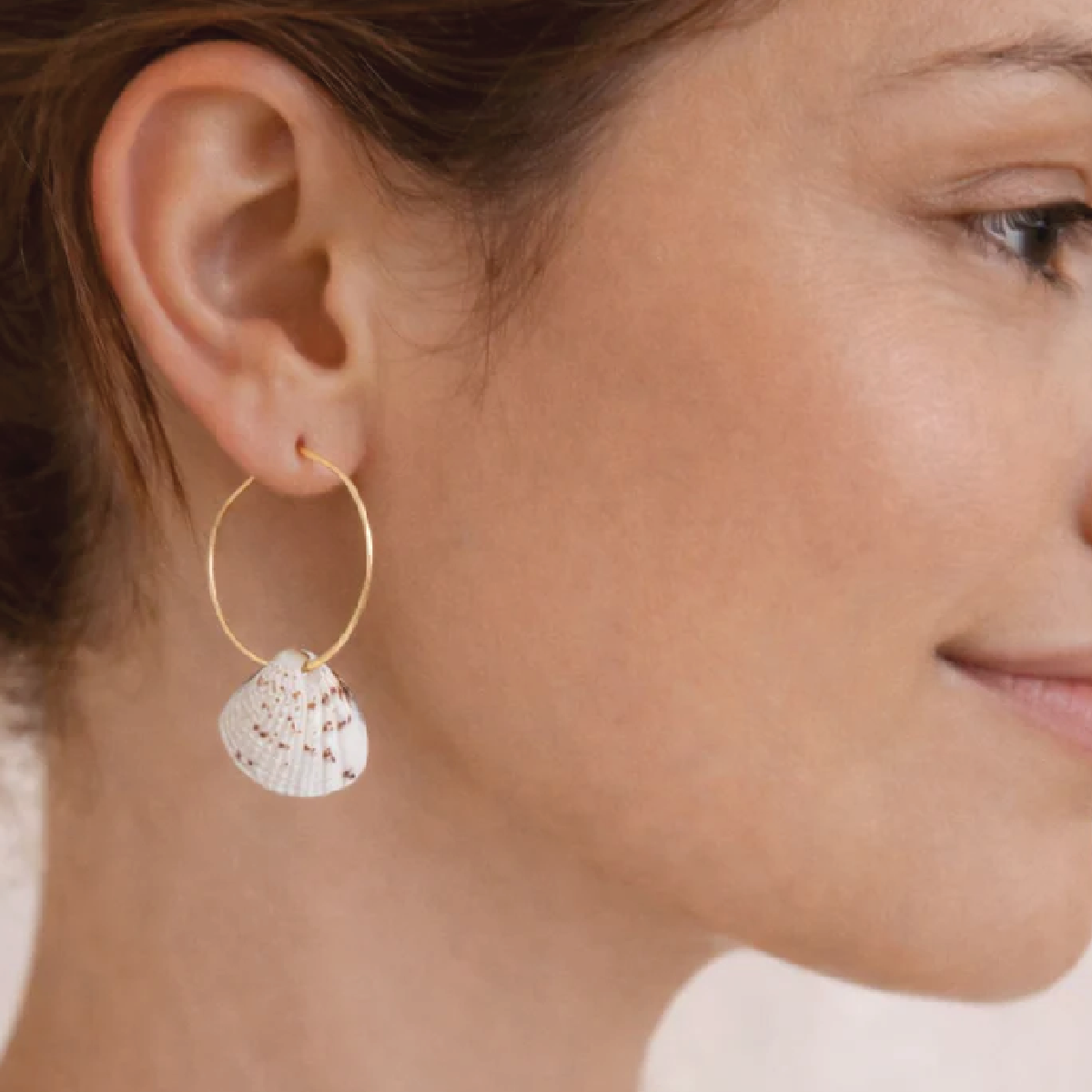 Close-up of a woman wearing a gold hoop earring with a white speckled shell pendant.